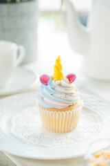 A whimsical unicorn cupcake with colorful frosting, sitting on a delicate doily. The cupcake is a delightful treat. A teapot and teacup sit near it.