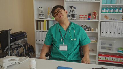 Young man in medical scrubs sits indoors in a clinic office, appearing stressed, with a stethoscope around his neck and a wheelchair in the background.