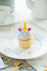 A single, beautifully decorated cupcake shaped like a unicorn. The cupcake features a colorful frosting design and a golden horn, presented on a white plate with tea accessories in the background