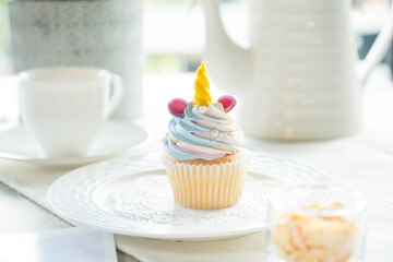 A charming unicorn cupcake on a plate, surrounded by elegant tableware. The cupcake features colorful frosting and a whimsical horn, set against a backdrop of soft light