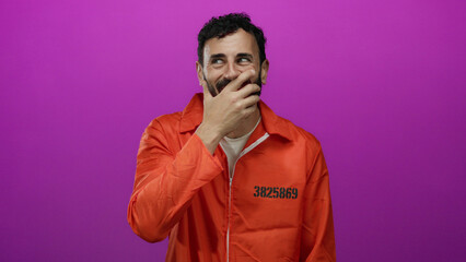 Hispanic man in an orange prison jumpsuit with a beard stands against a vibrant pink background, exuding a juxtaposition of confinement and light-heartedness while covering his smile.