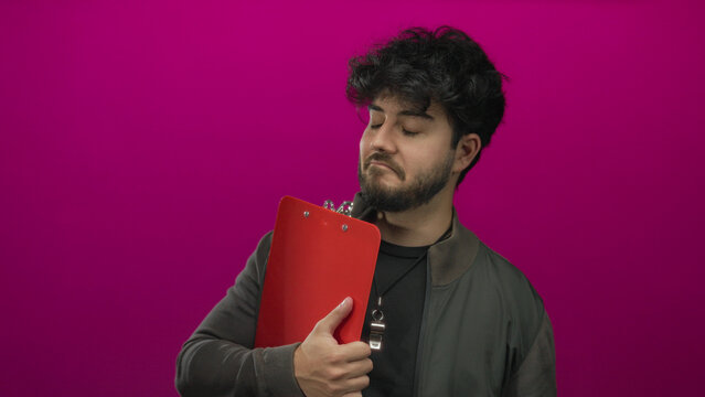 Young hispanic man with beard holding clipboard against pink background wearing jacket and whistle looking determined and thoughtful