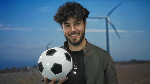 Young man with beard holding soccer ball in front of windmill in outdoor field setting with clear sky.