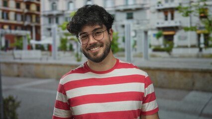 Young man with beard smiling outdoors on a street wearing glasses in a red striped shirt pointing happily on a bright day embracing fun and confidence with vibrant ambiance.