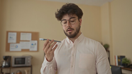 Young hispanic man in office records voice message using smartphone with focused expression and casual attire surrounded by notes and plants in organized indoor setting