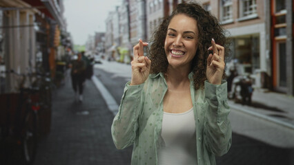 Young hispanic woman smiling with fingers crossed and hands pressed together in prayer on a city street lined with shops; hopeful.