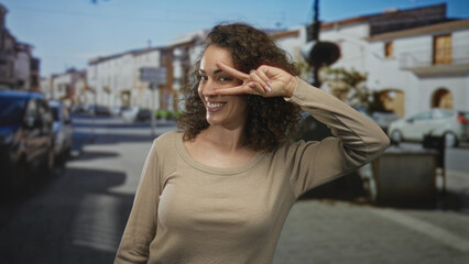 Fototapeta premium Woman holds two fingers in a v sign by her eye on a sunny urban street with parked cars and cobblestone pavement visible; playful curiosity.