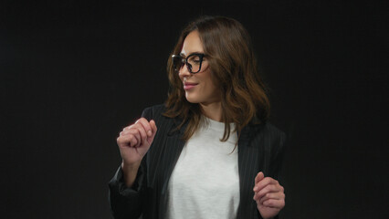 Woman dancing joyfully alone on isolated black background wearing glasses and stylish jacket, captured mid-movement with expressive gestures and a cheerful smile.