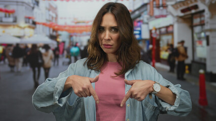 Woman pointing down on a busy city street with blurred pedestrians in the background, capturing an urban outdoor scene with banners overhead and a casual outfit.