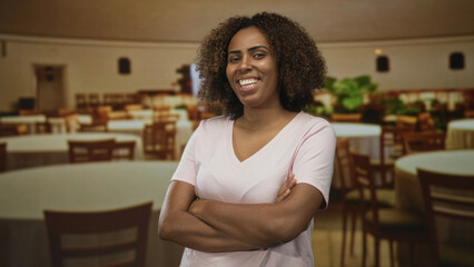 Woman with arms crossed smiling, bare forearms visible, wearing pink v neck shirt in a restaurant building interior with tables and chairs and potted plants; confidence hospitality.