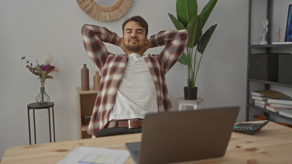 Young hispanic man working at laptop in modern office with plants and decor showcasing focus and concentration; dressed casually with plaid shirt and white t-shirt.