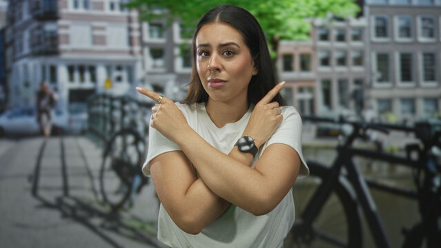 Woman wearing white t shirt with arms crossed x and index fingers pointing by canal railing on amsterdam street; defiant.