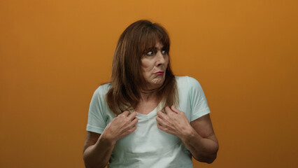 Fototapeta premium Hispanic senior woman in white shirt looks surprised against vibrant orange background, highlighting expressive emotion and mature facial features in an isolated setting.
