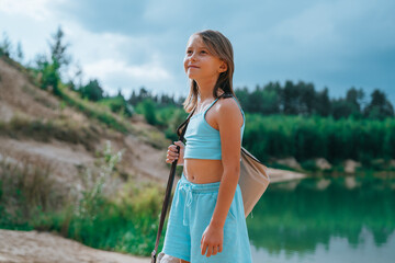 A young girl with a backpack gazes upward, smiling slightly under a cloudy sky, with lush greenery...