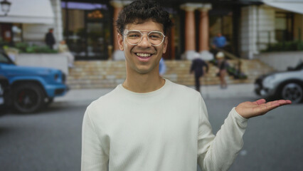 Fototapeta premium Man wearing white sweater and glasses smiling holds palm up on a busy city street near a building entrance; invitation.