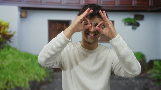 Man with smiling face makes hand binoculars to eyes in white sweater at building entrance; playfulness.