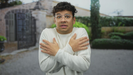 Fototapeta premium Young man hugs bare shoulders outside stone building entrance as he shivers in light sweater; anxiety.