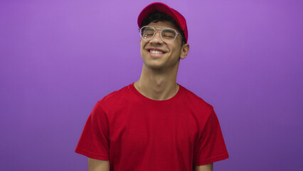 Obraz premium Youthful delivery man in red shirt and red cap with glasses smiles in studio against purple background; optimism.