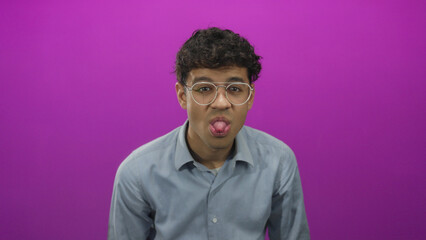 Young hispanic man wearing glasses sticks out tongue and holds hands by ears in a purple studio; playfulness.