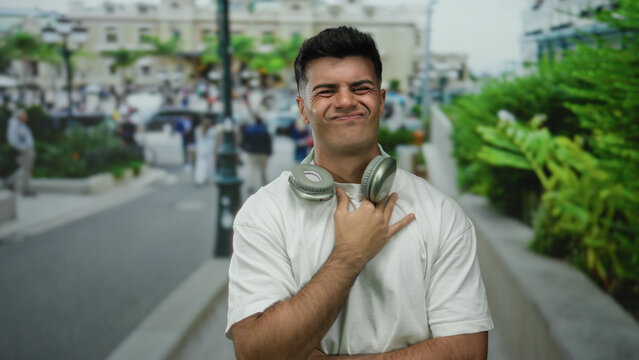 Young man with headphones around neck making expressions on city street with blurred background, wearing white shirt, showcasing a casual urban outdoor setting.