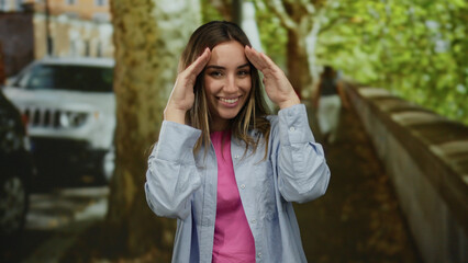 Fototapeta premium Young blonde woman smiling on a vibrant outdoor street with trees and cars, joyfully uncovering her face, exuding happiness and warmth in a candid city moment.