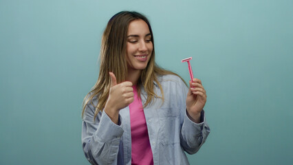 Fototapeta premium Young woman with blonde hair smiling and holding a pink razor against an isolated blue background while giving a thumbs up, showcasing confidence and style