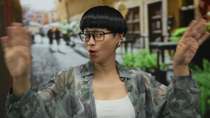 Woman smiling with short hair and glasses waves in busy city street with cafes and greenery...