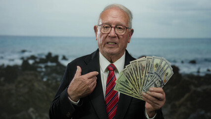 Senior man in business suit holding us dollars at seaside beach, conveying success and wealth with...