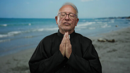 Elderly priest man in black robe praying peacefully on the seaside beach with waves and blue sky, embodying spirituality and serenity outdoors.