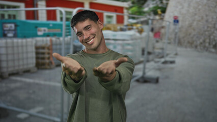 Naklejka premium Man extends arms in greeting wearing olive shirt at outdoor building site with stacked pallets and materials; invitation warmth.
