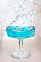 A festive cocktail in a crystal glass with blue ice on a bright white background with sparkles and glitter, copy space for text