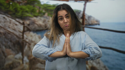 Hispanic woman with hands pressed in prayer on a seaside street railing near rocky cliffs; apology plea seeking forgiveness.
