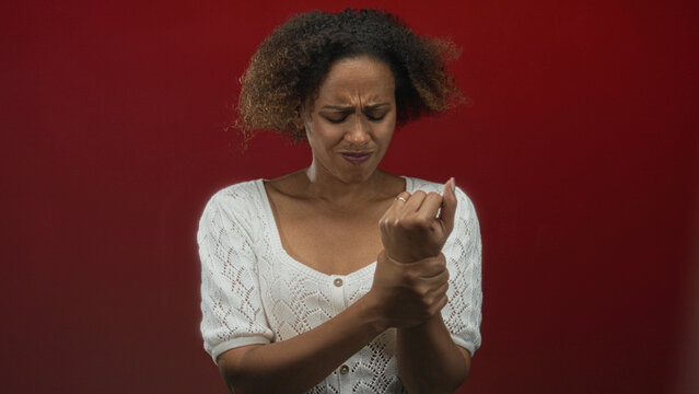 Woman grimacing while holding her wrist in red studio, wearing white knit sweater and visible ring, showing tense expression and tight grip; sprain distress.