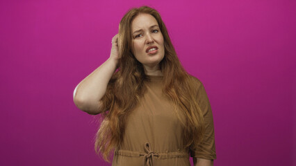 Woman with red wavy hair wearing brown dress scratches head and looks aside in pink studio; confusion.
