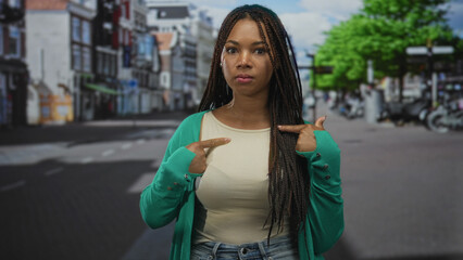 Woman points finger to chest on street, braided hair, green cardigan and tank top; self assertion pride.