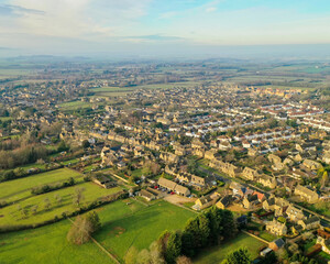 Aerial view of golden sunlight kissing the rooftops of quaint homes nestled among verdant fields, painting a serene landscape of rural charm, Cotswolds, England, United Kingdom.