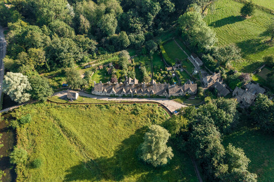 Aerial view of Arlington Row's honey-colored stone cottages nestling against verdant trees and fields under a soft, diffused light, Cotswolds, England, United Kingdom.