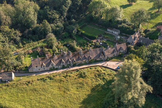 Aerial view of Arlington Row's honey-colored stone cottages nestled amidst lush greenery, a timeless scene of rural charm and architectural harmony, Cotswolds, England, United Kingdom.