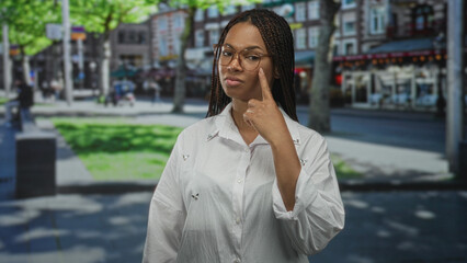 Fototapeta premium Woman pointing finger to eye on busy city street wearing glasses and white button shirt with braided hair, looking directly ahead; confidence.
