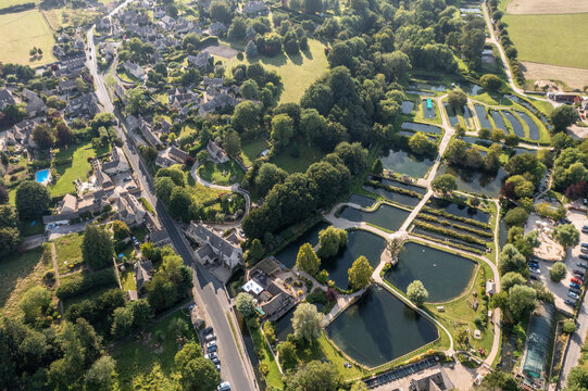 Aerial view of the Bibury Trout Farm's geometric ponds nestle beside a quaint village under a canopy of trees, a tapestry of nature and human design, Cotswolds, England, United Kingdom.