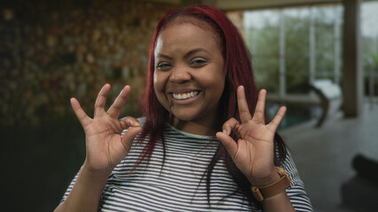 Woman with red hair smiling makes ok sign with both hands near pool inside a building spa area; approval ok.