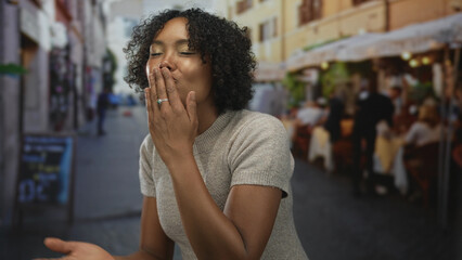 Fototapeta premium Woman blowing kiss with hand to lips on outdoor restaurant terrace overlooking blurred dinner tables and patrons; flirtation connection.