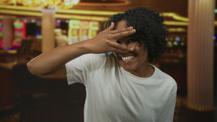 Woman covering eyes with hand and smiling behind fingers in casino building beside slot machines;...