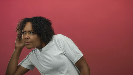 Young african american woman leans forward and holds hand to ear in pink studio listening pose; curiosity attention.
