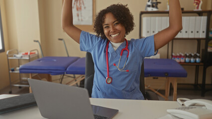 Woman nurse smiling raising arm over laptop on desk in clinic building; medical teamwork joy empowerment.