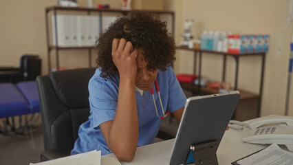 Woman wearing blue scrubs and stethoscope holding forehead and reading tablet at medical desk in building; stress.