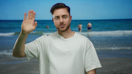 Young man with beard waving at the camera on a sunny beach with people in the background enjoying the seaside and the ocean waves under a clear blue sky, wearing casual white shirt.