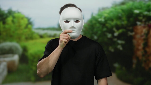 Young bearded man holding white mask in outdoor park setting with lush green trees visible in background creating an atmosphere of mystery and curiosity as he unveils his face to the camera