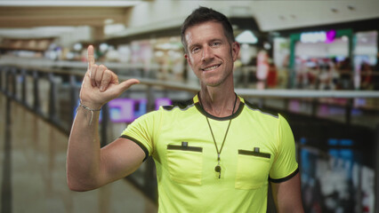 Fototapeta premium Man referee making shaka hand gesture with whistle lanyard and neon yellow shirt in a mall building; casual greeting friendly.