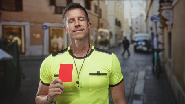 Man referee holds red card and whistle on a narrow street by a house entrance, eyes closed in a measured pause; calm authority.